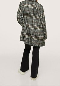 Cappotto a quadri in nero, bianco e marrone con taglio su misura, maniche lunghe e design della schiena dritto; abbinato a pantaloni neri svasati e sneaker.
