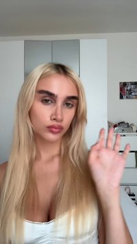 Langes, glattes blondes Haar mit einer geschmeidigen Textur, trägt ein weißes Top mit Spitzenakzenten, zeigt definierte Augenbrauen und einen neutralen Hintergrund.