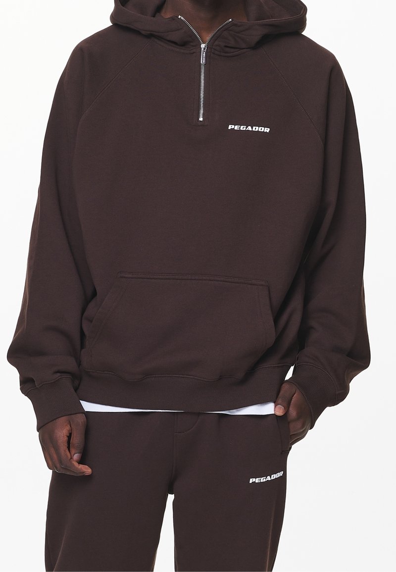 Personne portant un sweat à capuche marron foncé et un pantalon assorti avec des logos blancs "PEGADOR" et une fermeture éclair partielle à l'avant du sweat.
