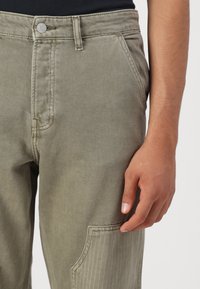 Olivgröna denimbyxor med en framficka, synlig söm och knappstängning. Strukturerad tyg med en något blekning utseende.