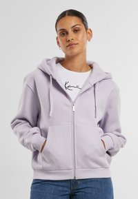 Felpa bianca con zip di colore viola chiaro, dotata di un ampio cappuccio, tasche frontali e polsini a costine. Indossata sopra una maglietta bianca con scritte nere.