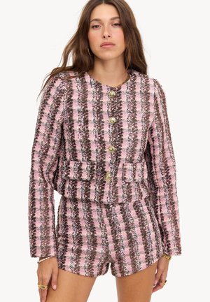 Mujer con chaqueta de tweed a rayas rosa, marrón y negra a juego con botones dorados y pantalones cortos de cintura alta.