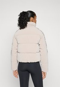Polstret jakke i lys beige med høy krage og teksturert stoff. Har et cropped design og side striper langs ermene.