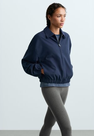 Femme portant une veste zippée bleu marine avec poignets et ourlet élastiques, debout les mains dans les poches, associée à des leggings gris.