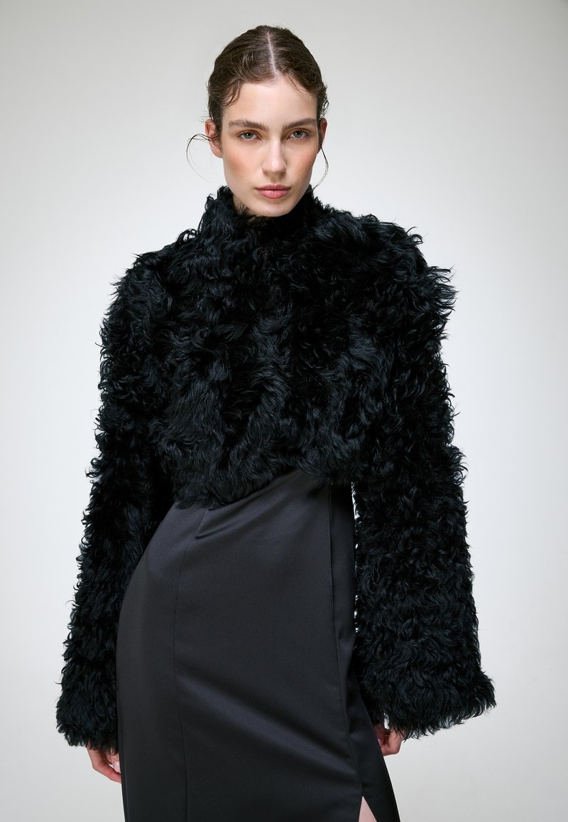 Femme aux cheveux plaqués en arrière porte une veste en fourrure texturée noire et une jupe en satin noire avec une fente latérale sur un fond uni.