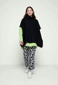 Poncho de malha preto oversized, sobre uma blusa de manga longa verde brilhante, combinado com calças com estampado floral em preto e branco e ténis brancos.