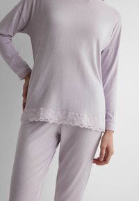 Frau trägt ein langärmliges Oberteil und Hosen in hellem Lila mit Spitzenbesatz an Saum und Bündchen, steht vor einer weißen Wand.