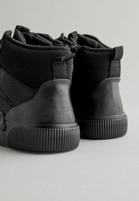 Schwarze Sneakers mit einem hohen Design. Hergestellt aus strukturiertem Stoff und Gummi, mit geriffelten Sohlen und einer Schlaufe für einfaches Anziehen.
