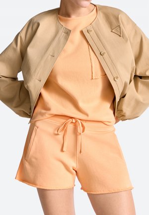 Naine kannab virsikuvärvi dressipluusi ja lühikesi pükse, millel on pael, ning beežikat kinnitatavat lühikest jopet.