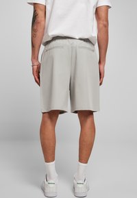 Uomo che indossa pantaloni corti grigi chiari, sneakers bianchi e calzini bianchi, in piedi con le spalle alla macchina fotografica su uno sfondo semplice.