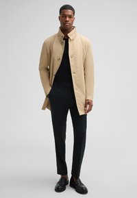Beige knoopjasje met hoge kraag, gedragen over een zwart shirt, gecombineerd met slim zwarte broek en zwarte schoenen. Soepele textuur en moderne pasvorm.