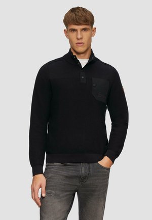 Zwarte gebreide trui met een kraag tot op een kwart, knoopsluiting en een borstzak. Heeft ribgebreide manchetten en zoom. Grijze denim jeans.