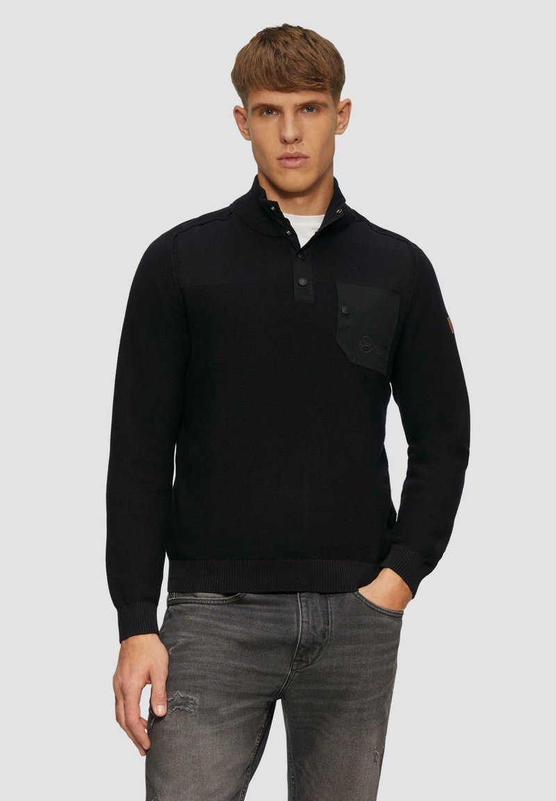 Schwarzer Strickpullover mit einem Kragen in Viertellänge, Knopfverschluss und einer Brusttasche. Verfügt über gerippte Bündchen und Saum. Graue Denim-Jeans.