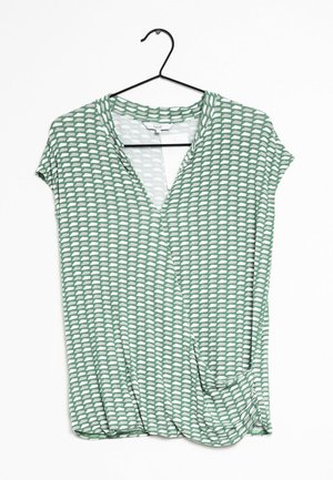 Haut vert à manches courtes avec un design croisé, décoré d'un motif abstrait blanc et d'un col en V. Tissu léger et lisse. Suspendu sur un crochet noir.