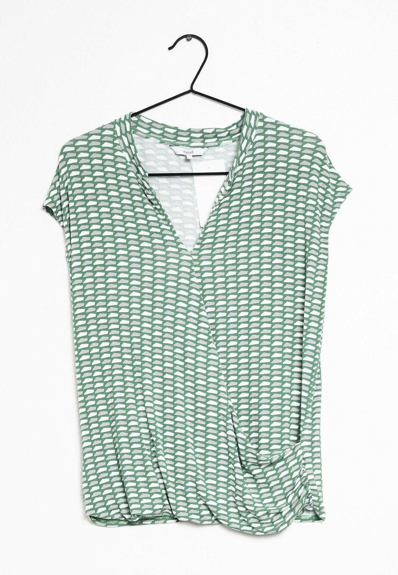 Haut vert à manches courtes avec un design croisé, décoré d'un motif abstrait blanc et d'un col en V. Tissu léger et lisse. Suspendu sur un crochet noir.