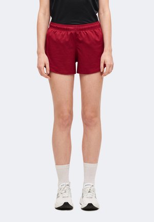 Personne portant un short de sport grenat, des chaussettes blanches, des baskets blanches et un haut noir, debout devant un fond clair uni.