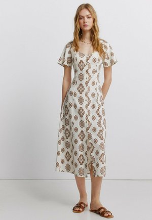 Femme portant une robe midi blanche avec des motifs géométriques marron, manches courtes, et des boutons sur le devant, associée à des sandales plates marron.