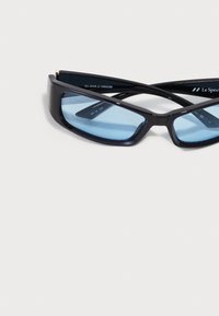 Ochelari de soare negri cu o ramă groasă și lentile cu nuanță albastră, având un design geometric și un branding subtil de-a lungul tâmplelor.