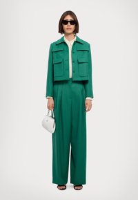 Green cropped jacket with two front pockets; matching wide-leg trousers; cream blouse underneath; holding a small white handbag; black sunglasses.