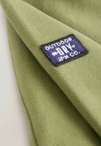 Tela verde texturizada con un pequeño parche rectangular azul marino etiquetado "OUTDOOR DRY JPN Co." cosido en la esquina.