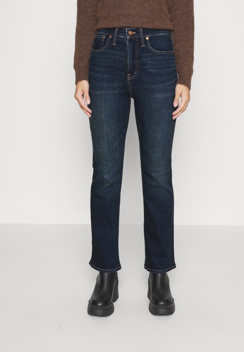 Personne portant un jean bleu foncé à jambes droites, un pull marron et des bottines noires, debout devant un fond blanc uni.