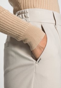 Beige žebrovaný svetr s dlouhými rukávy, který je spárován s krémovými kalhotami s boční kapsou a jemným brandingem poblíž kapsy.