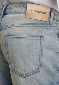 Poche arrière d'un jean en denim bleu clair avec une étiquette en cuir beige cousue au-dessus, affichant "2Y STUDIOS" en texte sombre.