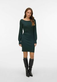 Žebrované pletené šaty tmavě zelené barvy s lodičkovým výstřihem, dlouhými rukávy a přiléhavým střihem, kombinované s černými kozačkami po kolena.