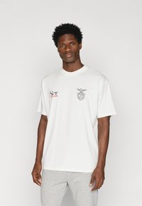 Biała bawełniana koszulka z krótkim rękawem, z czerwonym i czarnym napisem na klatce piersiowej oraz graficznym emblematem. Gładka tekstura, luźny krój.