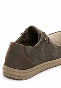 Zapato sin cordones de lienzo marrón con un diseño texturizado, que presenta un perfil bajo, suela de goma flexible y acentos laterales para facilitar su uso.