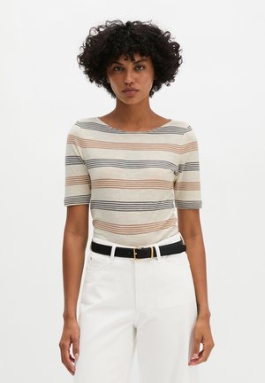 Frau mit kurzem lockigem Haar, die ein beiges Oberteil mit braunen und schwarzen horizontalen Streifen und weiße Hose mit schwarzem Gürtel trägt.