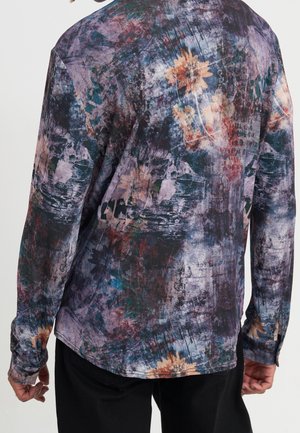 Overhemd met lange mouwen met abstracte, meerkleurige bloem- en textuurpatronen gedragen door een persoon die van zich af kijkt, gecombineerd met een zwarte broek.