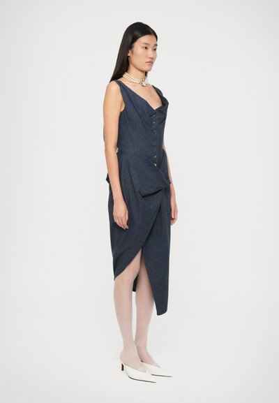 Mujer con un vestido asimétrico sin mangas de color azul oscuro con botones, collar de perlas, medias blancas traslúcidas y tacones puntiagudos blancos, de pie de perfil.