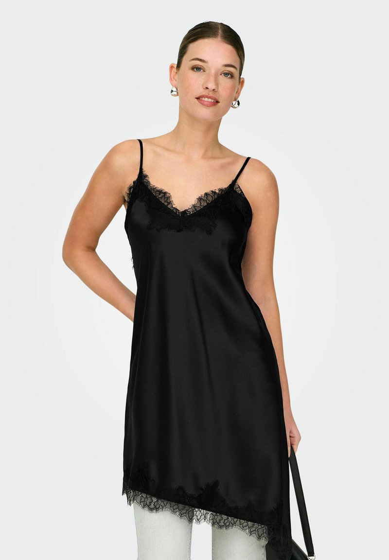 Femme portant une nuisette en satin noir avec une bordure en dentelle, associée à un pantalon blanc, debout avec une main sur la hanche et tenant une bretelle noire.