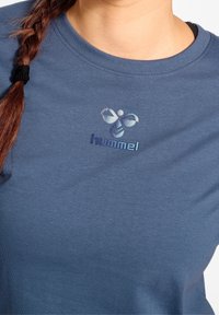 Niebieska bawełniana koszulka z okrągłym dekoltem oraz nadrukiem na przodzie z logo w postaci pszczoły i nazwą marki "hummel" w jasnoniebieskim kolorze.