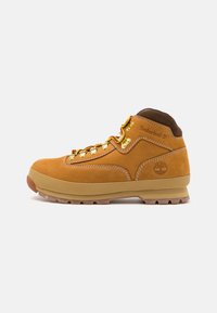 Botas de senderismo de cuero marrón con tobillo alto, cordones amarillos, ojales dorados y suela de goma con tacos para tracción. Logo de Timberland visible.