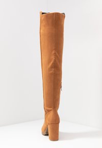 Brune ruskinds over-the-knee støvler med blokhæle, rund tå og lynlåsdetaillering på siden. Glat tekstur og tætsiddende design.