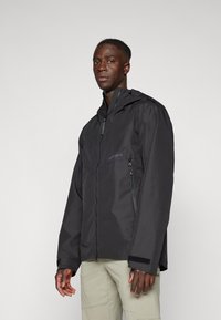 Veste noire imperméable avec fermeture éclair sur le devant, capuche réglable et poches latérales. Caractéristiques d'une texture lisse et d'un branding minimal.