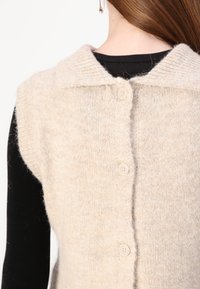 Gilet in maglia beige con collo a camicia, caratterizzato da una finitura testurizzata, design senza maniche e cinque bottoni chiari lungo la schiena.