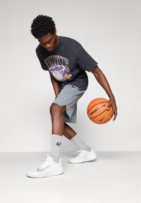 Basketbalový outfit Nike, ktorý pozostáva z čierneho grafického trička, svetlosivých šortiek, šedých ponožiek s logom a bielych športových topánok, driblírujúci s basketbalom.