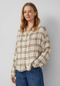 Frau mit lockigem, hellbraunem Haar, die ein beige-braunes karierter Hemd mit Knöpfen und blue Jeans trägt, steht mit einer Hand in der Tasche.