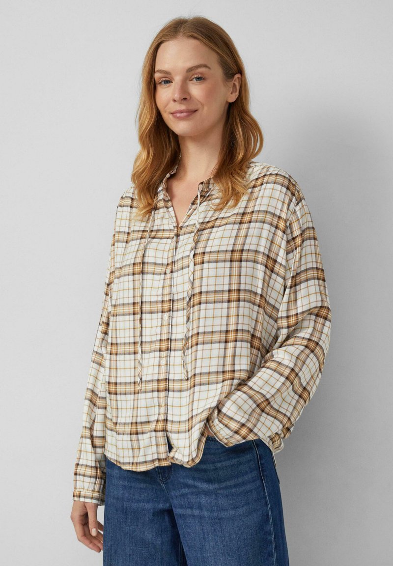 Frau mit lockigem, hellbraunem Haar, die ein beige-braunes karierter Hemd mit Knöpfen und blue Jeans trägt, steht mit einer Hand in der Tasche.