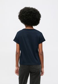 Άτομο με αφρο κουρέμα, φορώντας navy blue κοντομάνικο πουκάμισο και μαύρα τζιν, που στέκεται μπροστά σε λευκό φόντο.