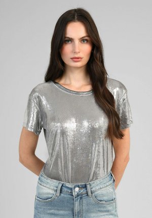 Femme aux longs cheveux foncés portant un haut court argenté brillant et un jean bleu clair, debout devant un fond gris uni.