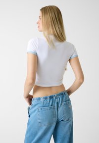 Weißes, kurz geschnittenes T-Shirt mit hellblauen Abschlüssen an den kurzen Ärmeln, kombiniert mit hellblauen Denim-Hosen mit elastischem Bund und zwei Gesäßtaschen.