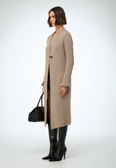 Femeie stând îmbrăcată într-un cardigan lung bej tricotat, cizme negre din piele, ținând o geantă mare neagră, pe fundal simplu și deschis la culoare.