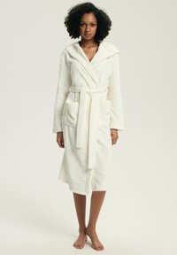 Kobieta stojąca boso, ubrana w biały, z kapturem, sięgający kolan szlafrok kąpielowy z paskiem i przednimi kieszeniami na jednolitym tle.