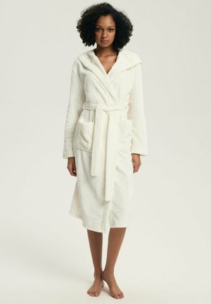 Kobieta stojąca boso, ubrana w biały, z kapturem, sięgający kolan szlafrok kąpielowy z paskiem i przednimi kieszeniami na jednolitym tle.