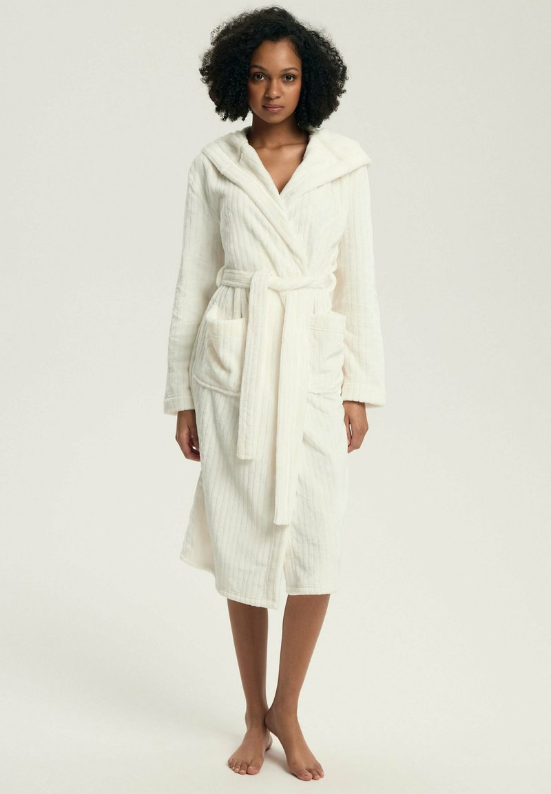 Kobieta stojąca boso, ubrana w biały, z kapturem, sięgający kolan szlafrok kąpielowy z paskiem i przednimi kieszeniami na jednolitym tle.