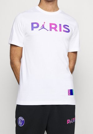 Homme portant un t-shirt blanc avec le texte "PARIS" en violet-rose et le logo Jumpman, associé à un pantalon noir arborant les logos du Paris Saint-Germain.